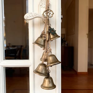 Może przedstawiać: Dekoracyjna zawieszka z antycznymi mosiężnymi dzwonkami, kluczami i urokiem w kształcie słońca. Dzwonki zawieszone są na naturalnej lince, tworząc rustykalną i vintage estetykę. Układ przymocowany jest do białej klamki.