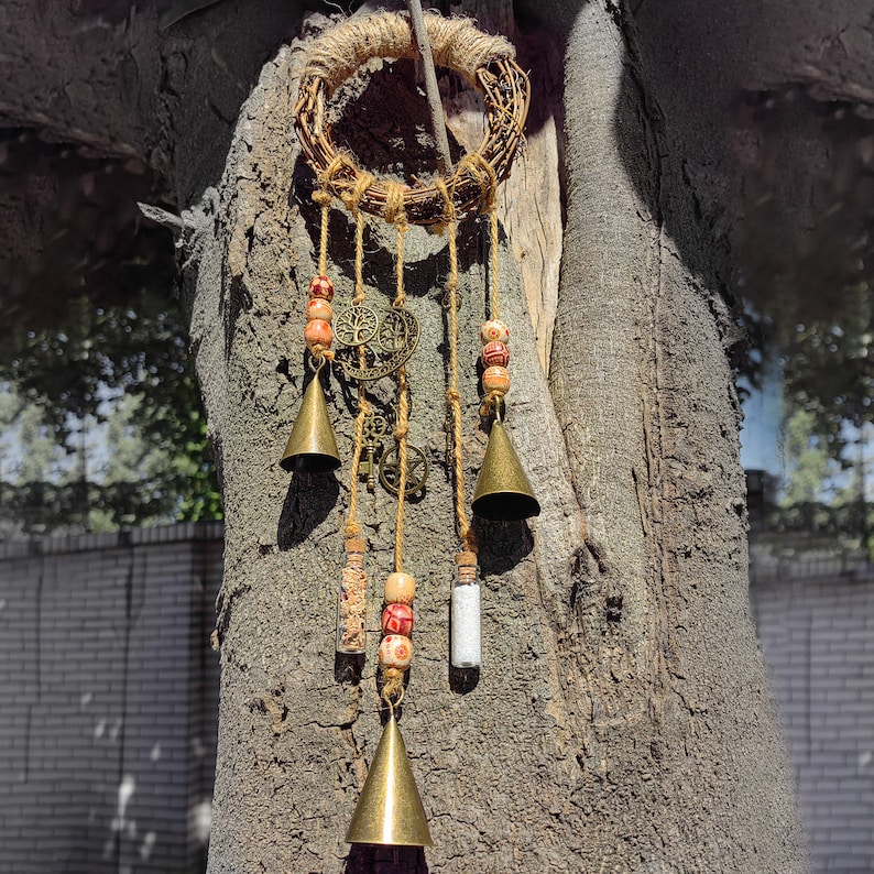 May include: A brown twig wreath with a beaded and bell wind chime hanging from it. The chime features small glass bottles, beads, and bells. The wreath is hanging on a tree trunk.