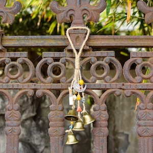 Op de afbeelding: Een set gouden bellen die aan een robuust metalen hek hangen. De bellen zijn bevestigd aan een touwtje met kralen en bedels, waaronder een halve maan bedel.