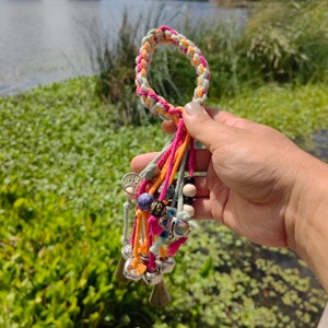 以下が含まれることがあります： ピンク、オレンジ、グリーンの糸で編まれたカラフルなブレスレット。ベル、ビーズ、平和のサインのチャームなど、さまざまなチャームが付いています。