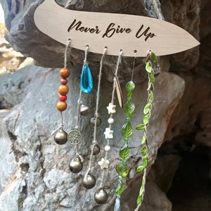 Puede incluir: Carillón de viento de madera con las palabras "Never Give Up" grabadas en la parte superior. El carillón presenta una variedad de cuentas, campanas y encantos, incluyendo hojas verdes, cuentas de vidrio azul y verde, estrellas blancas y un pequeño encanto de árbol.