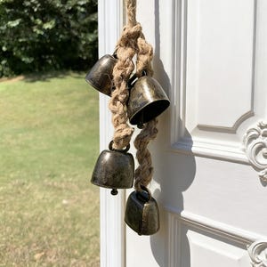 Puede incluir: Un colgante decorativo de cuatro campanas de color bronce suspendidas de una gruesa cuerda trenzada. Las campanas tienen un aspecto rústico y envejecido y están unidas a la cuerda con pequeños lazos metálicos. La cuerda está anudada en la parte superior.