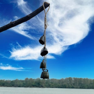 Könnte beinhalten: Ein Satz von vier kleinen, dunkelbraunen Glocken, die an einer Bindfadenkette gegen einen blauen Himmel mit weißen Wolken hängen. Die Glocken sind mit Knoten zusammengebunden und hängen an einem Ast.