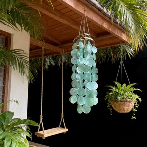 May include: A hanging wind chime with light green, circular accents suspended from a gold-colored ring. A wooden swing hangs from the roof, and a hanging basket with green plants is also visible. The scene is set outdoors.