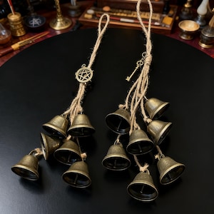May include: Two sets of bronze-colored bells hanging from natural twine. One set features a pentagram charm, the other a key charm. Each set has multiple small bells, suitable for decorative or ritual use.