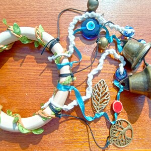 May include: A white wooden ring with green leaves wrapped around it, decorated with blue and white evil eye charms, gold bells, and a gold leaf charm. The ring is hanging from a white cord with blue ribbon and black string.