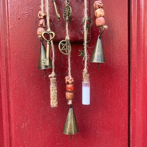 May include: A macrame wall hanging with bells, keys, and small glass vials. The macrame is made with natural twine and features wooden beads with a red and white pattern.