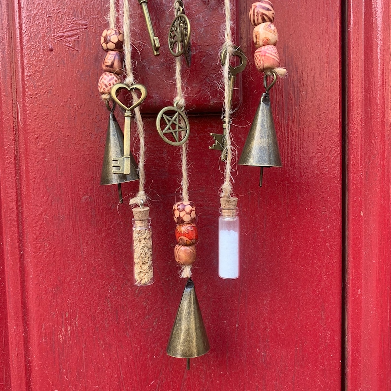 May include: A macrame wall hanging with bells, keys, and small glass vials. The macrame is made with natural twine and features wooden beads with a red and white pattern.