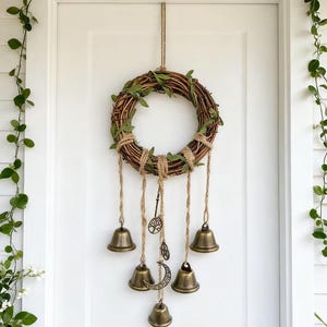 May include: A rustic wreath made of woven branches, adorned with green leaves and twine. Six bronze bells hang from the wreath, along with decorative charms. The wreath is suspended on a white door, with greenery on either side.