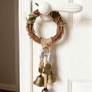 May include: A handcrafted door wreath made of woven brown twigs, adorned with green leaves and jute twine. It features a hanging strand with blue beads, a metal ghost charm, and three brass bells. The wreath hangs from a white door handle.