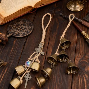 Puede incluir: Dos campanillas de viento de color bronce con colgantes de cuerda de yute. Una campanilla presenta una silueta de bruja, una cruz y un cristal. La otra tiene cinco campanas. Un libro abierto, una vela y otros objetos místicos están en el fondo.
