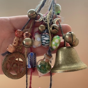 May include: A collection of charms and beads hanging from strings, including bells, wooden beads, glass beads, and a small glass bottle filled with green foliage. The charms are tied together with twine and feature a variety of colors, including green, red, blue, and white.