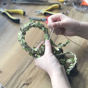 May include: A person is tying a piece of twine around a wreath made of woven twine and green leaves.
