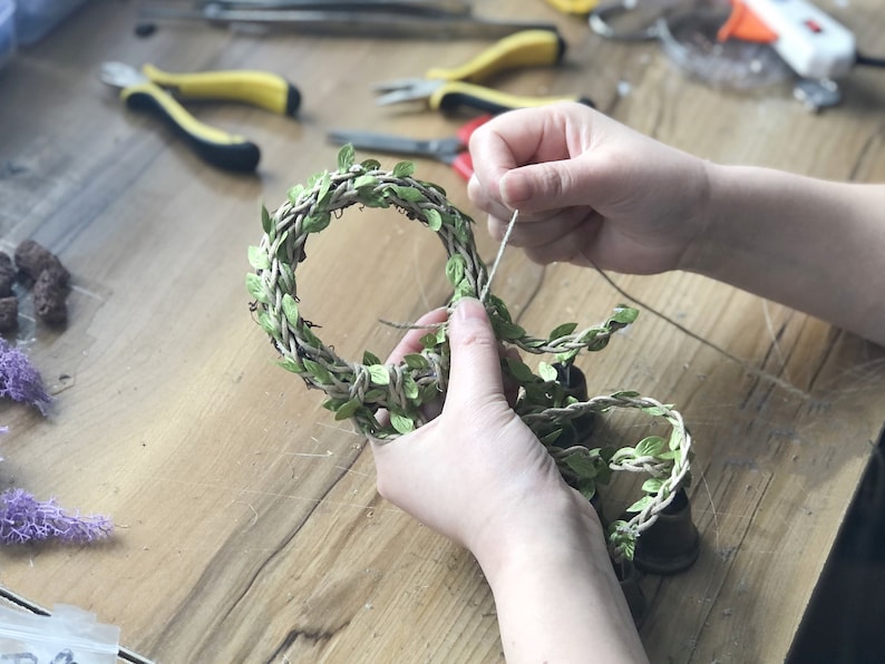 May include: A person is tying a piece of twine around a wreath made of woven twine and green leaves.