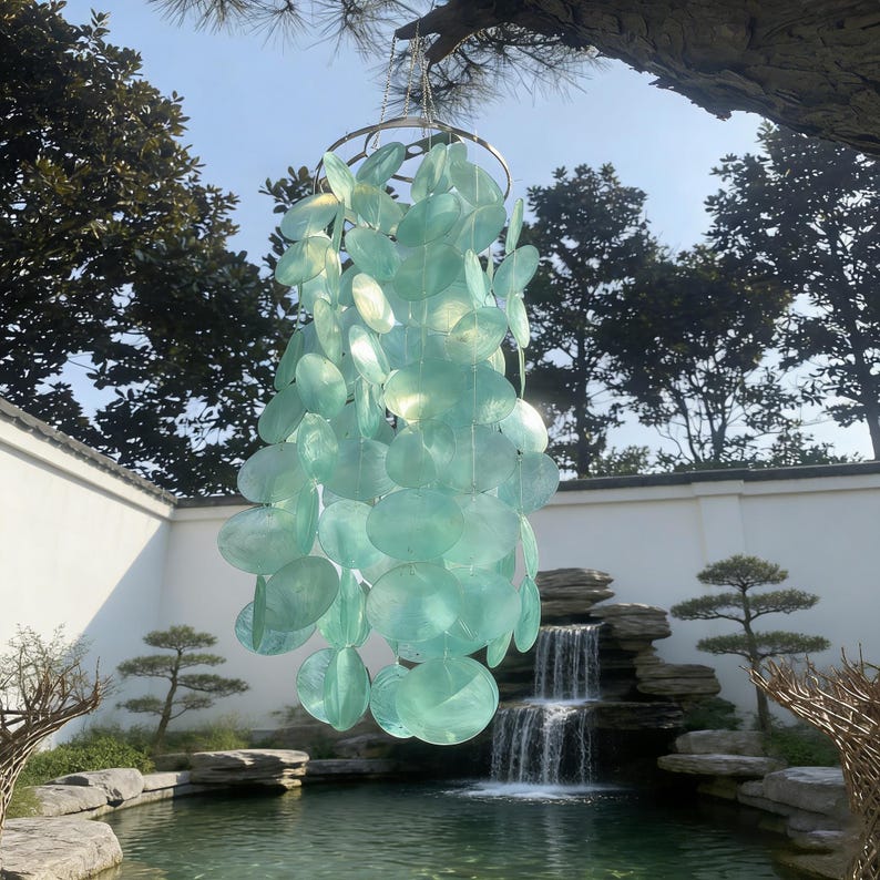 May include: A hanging wind chime with a silver metal ring and strands of translucent, light-blue, oval-shaped pieces. The chime is suspended from a tree branch against a backdrop of a garden with a pond and waterfall.
