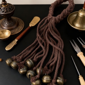 Peut inclure: Une corde marron avec plusieurs brins, chacun se terminant par une petite cloche en laiton antique. Les cloches ont une forme classique et sont attachées à la corde avec des glands. La corde est nouée en haut, créant une boucle.