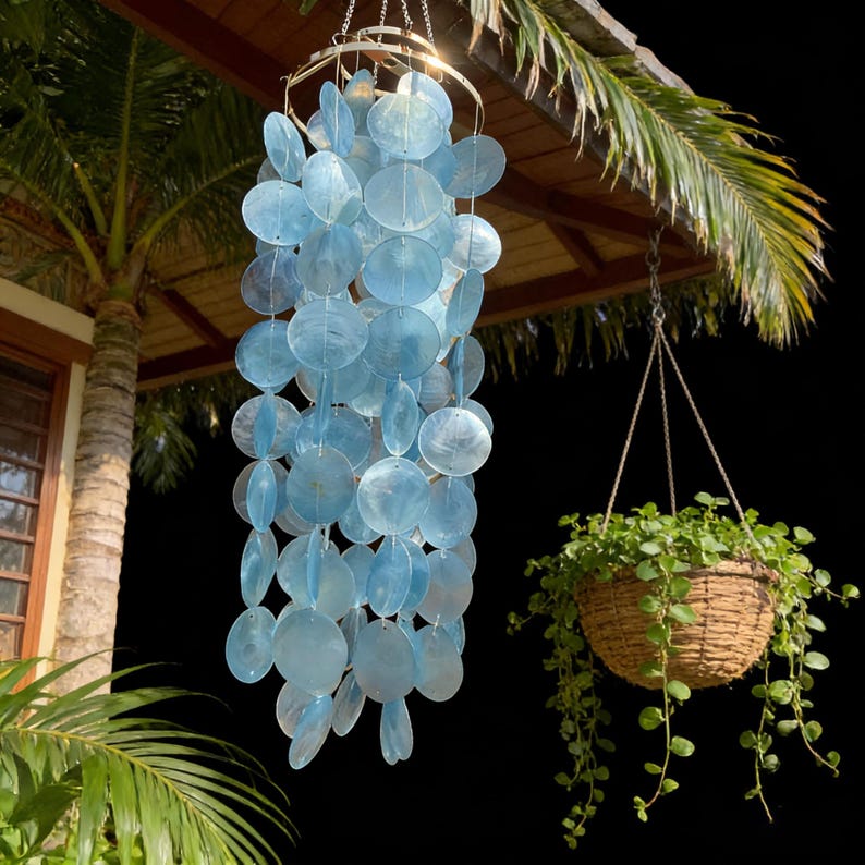 May include: A hanging wind chime with light blue, round, shell-like discs. The chime is suspended from a metal ring and hangs below a wooden roof. A hanging basket with green plants is also visible.