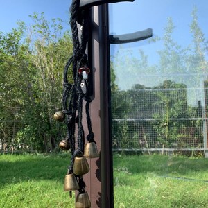 以下が含まれることがあります： 小さなアンティーク真鍮のベルがいくつかぶら下がっている装飾的な黒いロープ。キノコ型の小さなチャームがロープに取り付けられています。ロープは、黒いハンドルが付いた茶色のドアフレームにぶら下がっており、緑の草の背景があります。