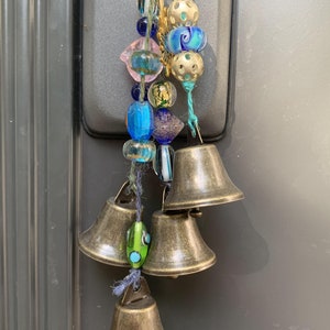 May include: A set of four brass bells with a string of colorful glass beads and gold accents. The bells are hanging from a door handle.