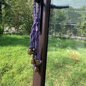 以下が含まれることがあります： 小さな真鍮のベルがいくつかぶら下がっている紫色の編みロープ。ロープはドアノブに取り付けられています。背景には緑の草と葉が見えます。