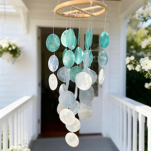 Puede incluir: Un carillón de viento colgante con un anillo de madera natural y cuerdas de discos iridiscentes, redondos, aguamarina y blancos en forma de concha. El carillón de viento cuelga frente a un porche blanco con flores blancas.