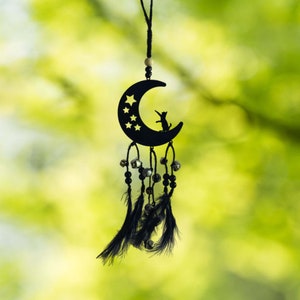 Puede incluir: Atrapasueños de metal negro con una luna creciente con estrellas y una silueta de gato. El atrapasueños tiene plumas negras, cuentas y pequeñas campanas colgando debajo. El fondo es verde borroso.