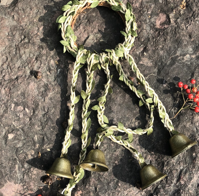 May include: A wreath made of green leaves and twine with four small brass bells hanging from it.