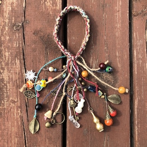 Puede incluir: Un brazalete trenzado multicolor con una variedad de encantos, incluyendo cuentas, hojas, campanas y una pequeña calabaza. El brazalete está hecho con fibras naturales y tiene un estilo rústico y bohemio.