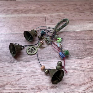 May include: A handmade wind chime with four brass bells, a wooden bead, and a variety of colorful beads and charms. The chime is suspended from a brown cord.