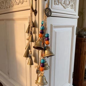 May include: A collection of brass bells hangs from a natural rope, adorned with colorful beads. The bells are of varying sizes and shapes, creating a rustic and bohemian aesthetic. The bells are hanging on a white cabinet with ornate carvings.