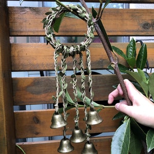 May include: A green leaf wreath with five gold bells hanging from it. The wreath is made of woven twine and leaves.