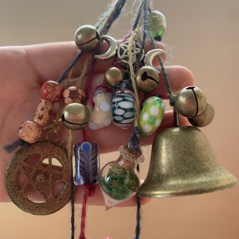 May include: A collection of charms and beads hanging from strings, including bells, wooden beads, glass beads, and a small glass bottle filled with green foliage. The charms are tied together with twine and feature a variety of colors, including green, red, blue, and white.