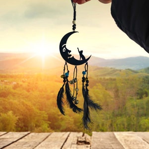 Puede incluir: Atrapasueños negro con una luna creciente y una silueta de gato, colgando de una cuerda con plumas negras y cuentas. El atrapasueños está frente a un fondo borroso de una puesta de sol y árboles.