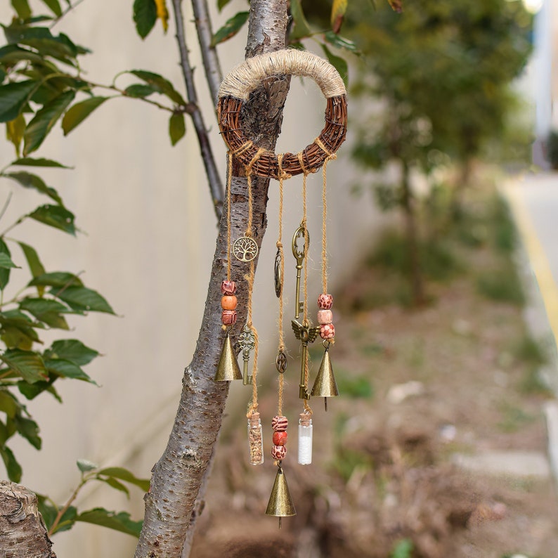 May include: A rustic-style wind chime with a woven twig ring, hanging from a tree branch. The chime features multiple strands with small bells, keys, beads, and a charm with a tree of life design.