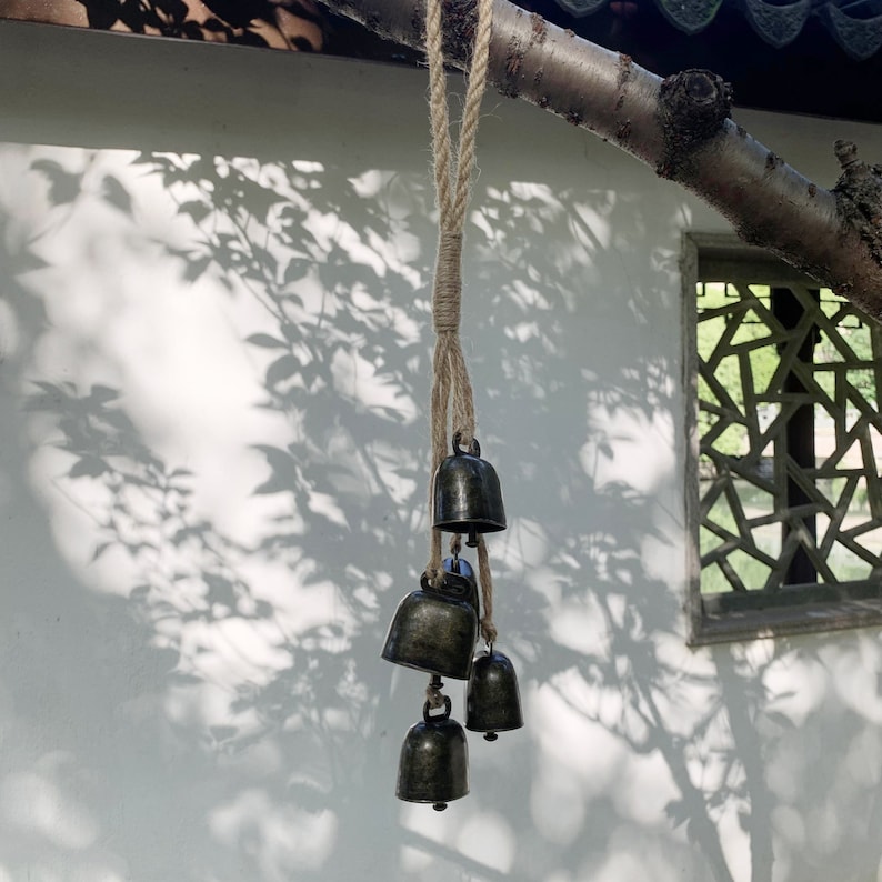 May include: A set of five small, antique-looking bells hanging from a length of twine. The bells are a dark bronze color and have a slightly aged appearance. The twine is a natural beige color and is tied to a branch above the bells.