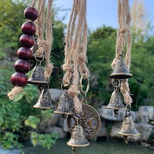 May include: A hanging wind chime featuring small bronze bells and dark red wooden beads strung on natural jute rope. The bells have decorative patterns and are suspended near a bronze pendant with a crescent moon design.