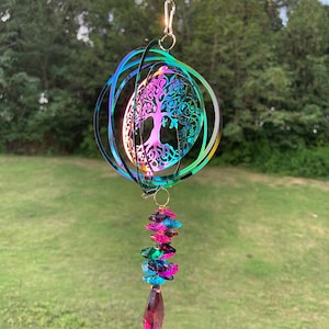 Puede incluir: Un adorno giratorio decorativo con un diseño de árbol de la vida. El adorno giratorio tiene anillos iridiscentes y un colgante de cristal colorido. El árbol de la vida está recortado del centro del adorno giratorio. El colgante está hecho de cristales coloridos.