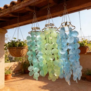 May include: Three hanging wind chimes with a brass-colored metal frame and chains. The chimes feature iridescent, circular, shell-like pieces in shades of green and blue. Wicker baskets with plants hang nearby.