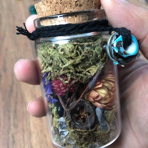 May include: A small glass jar with a cork lid, filled with dried flowers, moss, and crystals. The jar is tied with a black string and has a blue and white glass bead attached.