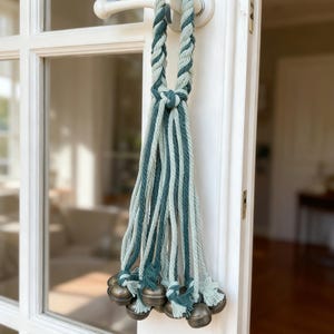 May include: A decorative door hanger featuring a braided rope in shades of teal and seafoam green. The rope is knotted and adorned with multiple strands of teal and seafoam green rope, each ending with a small, antique-style bell. The door hanger is attached to a white door.
