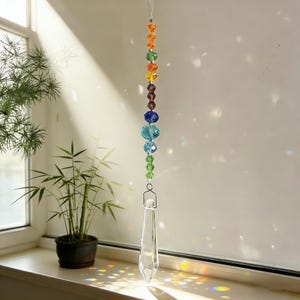 Può includere: Un acchiappasole di cristallo sospeso con una serie di perline colorate arcobaleno. Le perline sono disposte verticalmente, passando dall'arancione al viola, con un prisma di cristallo trasparente in basso. Sospeso da un filo sottile.