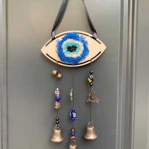 Peut inclure: Un carillon en bois en forme d'œil avec un motif œil mauvais bleu et blanc, des perles bleues et noires et des cloches dorées. Le mauvais œil est entouré de fil bleu et blanc.