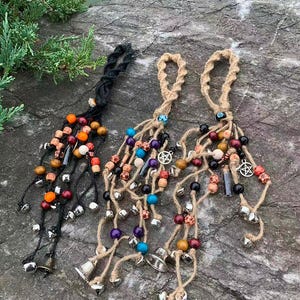 以下が含まれることがあります： ジュートコード、木製ビーズ、小さな銀色のベルが付いた、手作りのウィンドチャイムが3つ。ビーズはオレンジ、赤、黒、紫など様々な色があります。各チャイムの上部には吊り下げ用のループが付いています。