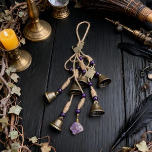 May include: A handcrafted hanging charm featuring small brass bells, purple beads, and a raw amethyst crystal. The charm is suspended from a natural twine loop and adorned with metal cat charms. The arrangement is set against a dark wooden surface.