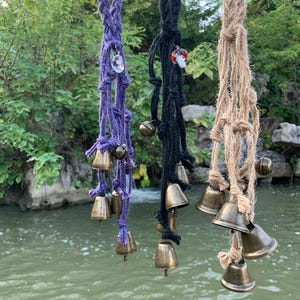 以下が含まれることがあります： 紫、黒、ナチュラルカラーの三色の編みロープに真鍮のベルが付いた、吊り下げ式のウィンドチャイム。各チャイムにはユニークなチャームが付いています。背景には水面と緑豊かな木々が写っています。