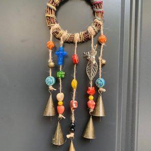 Puede incluir: Una pequeña corona tejida con cuentas de colores y campanas colgando de ella. Las cuentas son rojas, naranjas, amarillas, verdes, azules y blancas. Las campanas son doradas y tienen un aspecto rústico. La corona es de color marrón claro y tiene un aspecto natural y rústico.