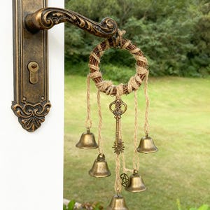 May include: A decorative door knocker with a rustic aesthetic. It features a small woven wreath with hanging bronze bells and a key charm. The door handle is ornate, with a bronze finish, and is mounted on a white door.