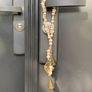 Pode incluir: Uma guirlanda de contas de madeira com contas em forma de estrela penduradas em uma maçaneta de porta. A guirlanda tem três pequenos sinos dourados presos à parte inferior.