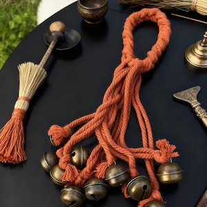 May include: A collection of spiritual tools on a black surface. The central item is a burnt orange rope with multiple strands, each ending in a small, antique brass bell. Other items include a small broom, a bowl, and a metal tool.