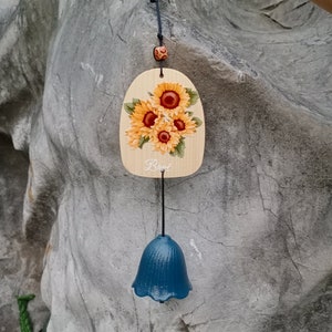 Puede incluir: Un carillón de viento de madera con una campana azul y un diseño de girasol pintado. La pieza de madera tiene la palabra "Burst" escrita en ella.